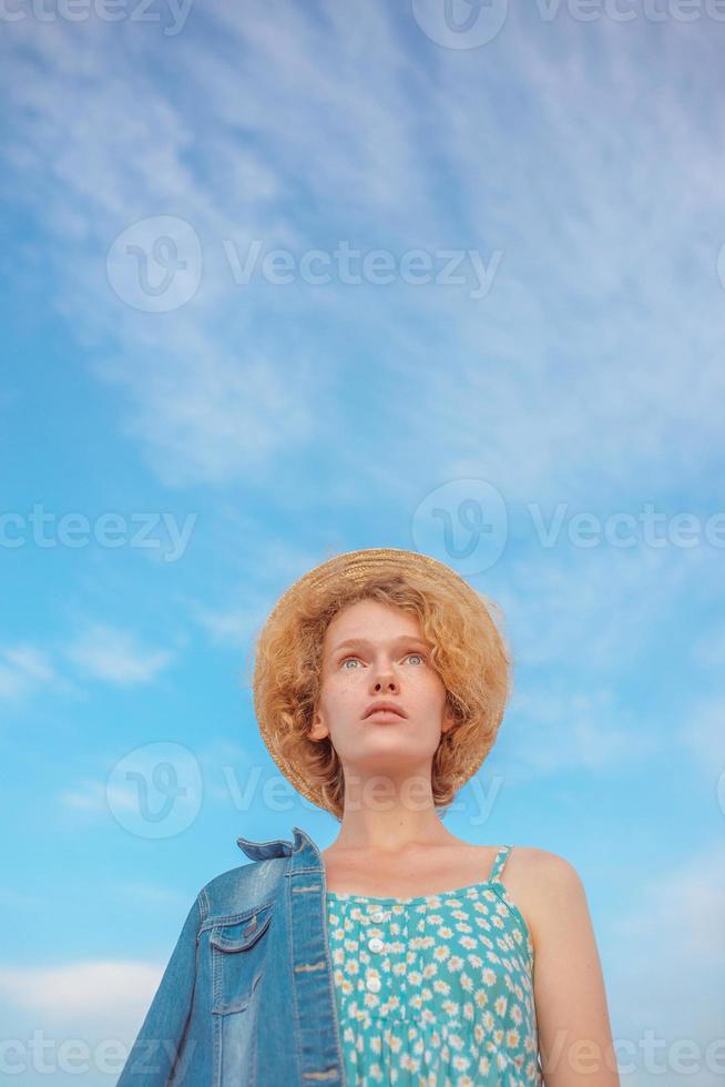 young curly redhead woman in straw hat, blue sundress and jeans jacket standing on blue sky background. Fun, summer, fashion, shooting, travel, youth concept. Copy spase photo