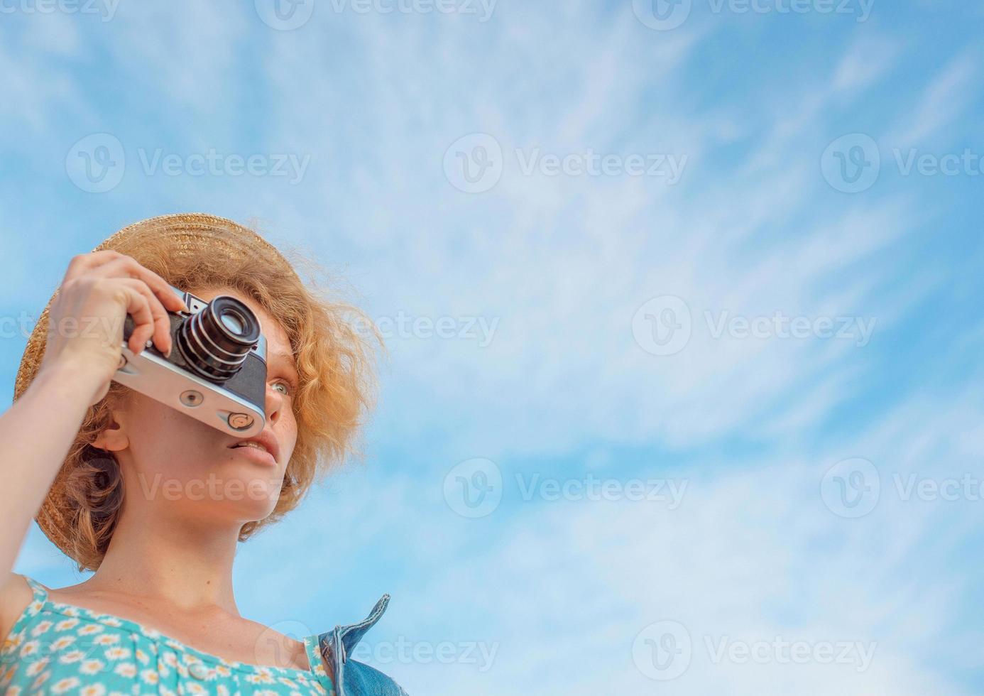 young curly redhead woman in straw hat, blue sundress and jeans jacket standing with vintage camera and taking pictures on blue sky background. Fun, summer, fashion, shooting, travel, youth concept photo