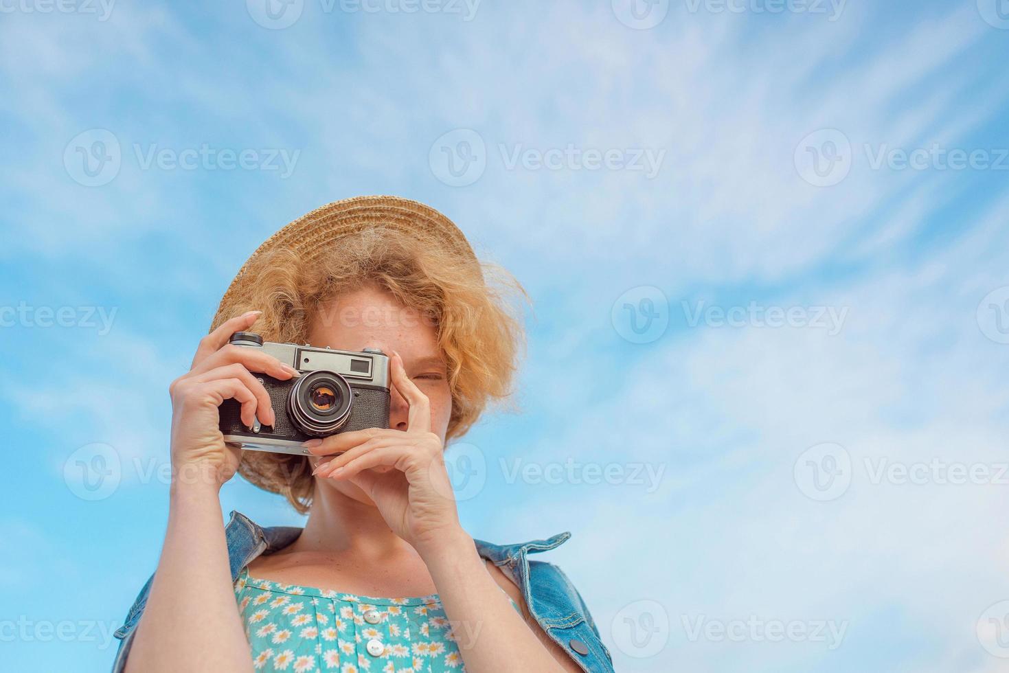 young curly redhead woman in straw hat, blue sundress and jeans jacket standing with vintage camera and taking pictures on blue sky background. Fun, summer, fashion, shooting, travel, youth concept photo