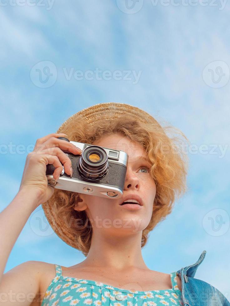 young curly redhead woman in straw hat, blue sundress and jeans jacket standing with vintage camera and taking pictures on blue sky background. Fun, summer, fashion, shooting, travel, youth concept photo