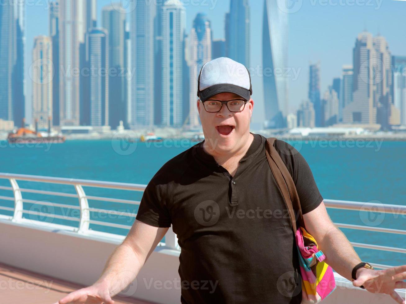 portrait of smiling Europian caucasian redhead tourist man in glasses and hat Impressed in modern Dubai, United Arabic Emirates on the cityscape, skylines and sea background in sunny day photo