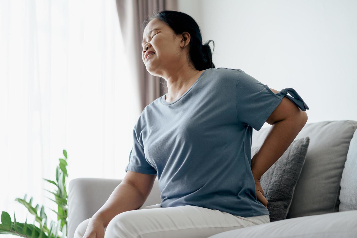 Adult Asian woman is sitting on the sofa and holding her lower back suffering from injured back. Health care and back pain concept. photo