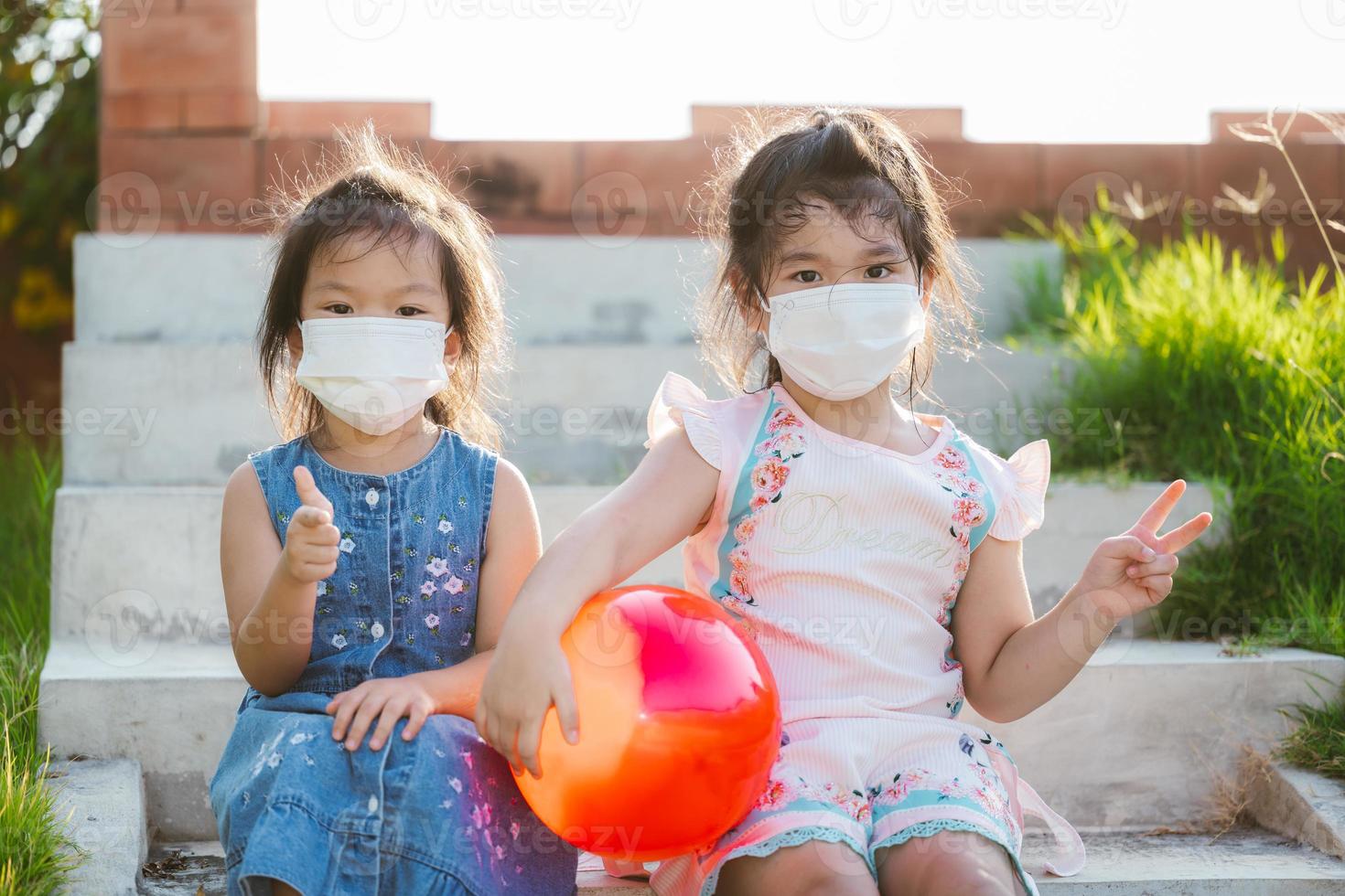 Children wearing white face mask are sitting on steps. Child holding red ball. Kid holds up finger to V sign. Beautiful girl aged 5 year old. photo