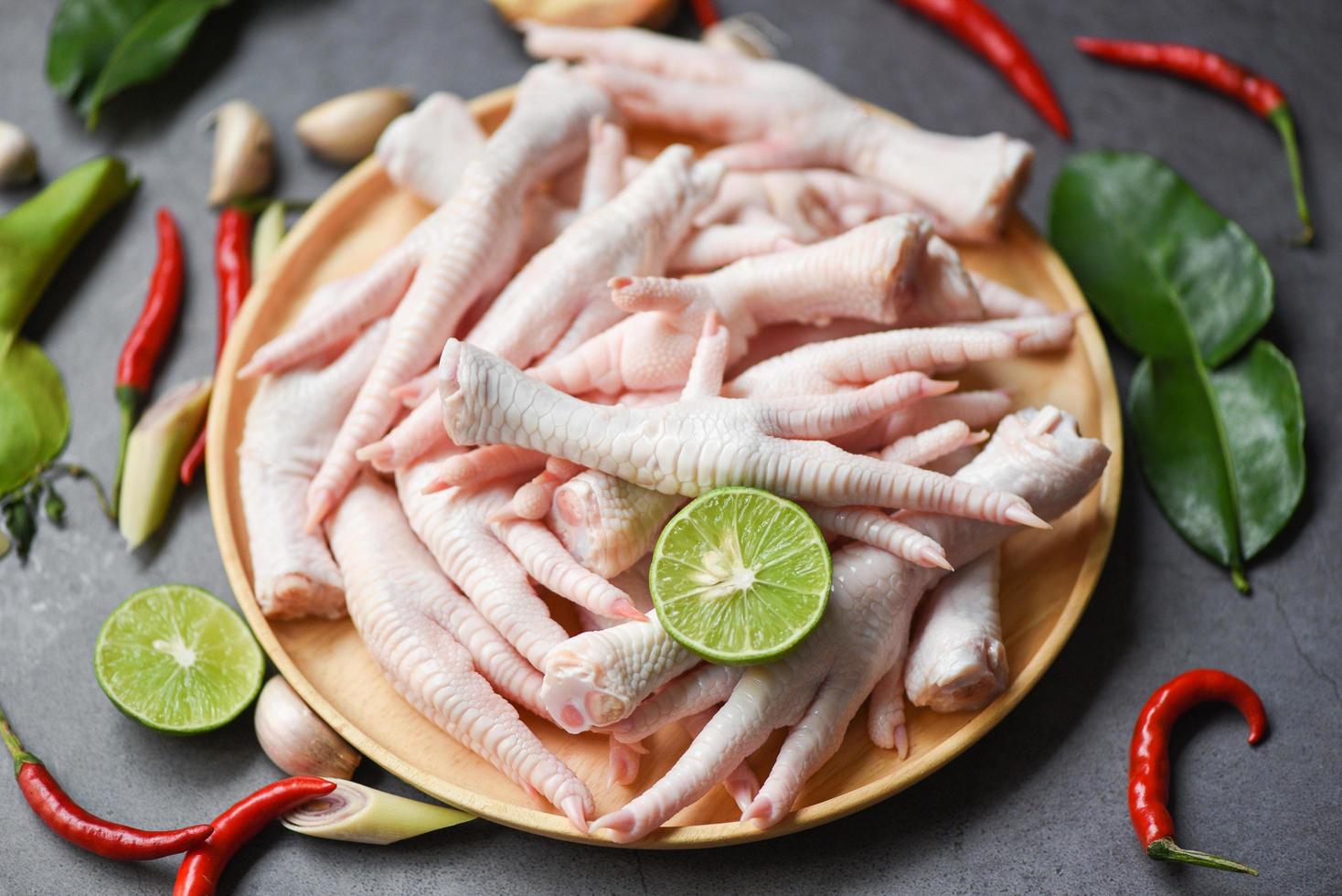 patas de pollo en un plato de madera con hierbas y especias limón chile ajo hojas de lima kaffir, patas de pollo crudas frescas para sopa de comida cocinada en el fondo oscuro de la cocina foto