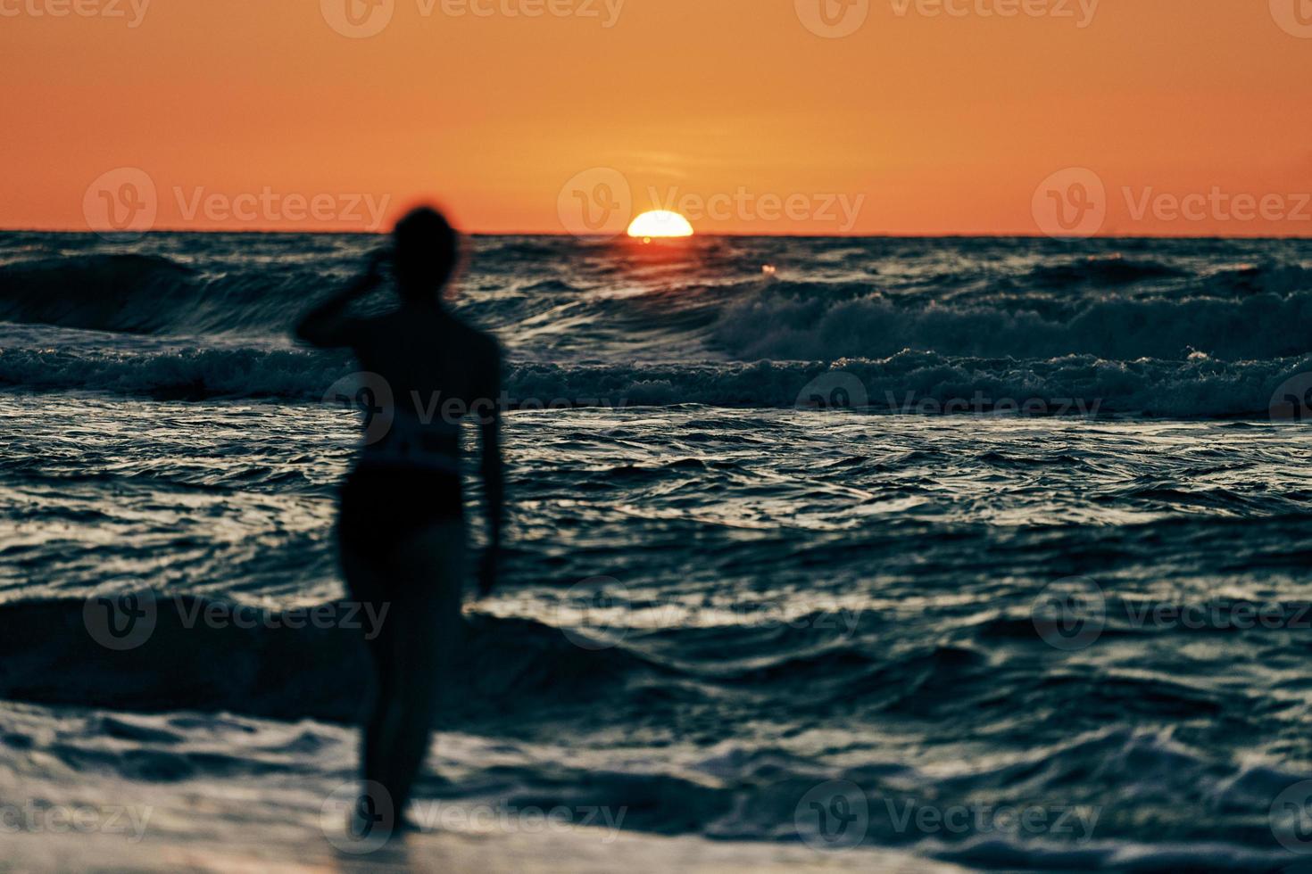 Female silhouette in blue sea waves at summer sunset, half sun below horizon, beachfront holiday photo