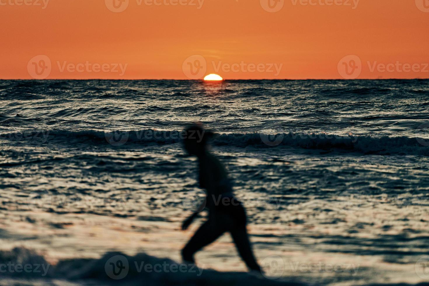 Female silhouette walking in sea waves at summer sunset, half sun below horizon, beachfront holiday photo