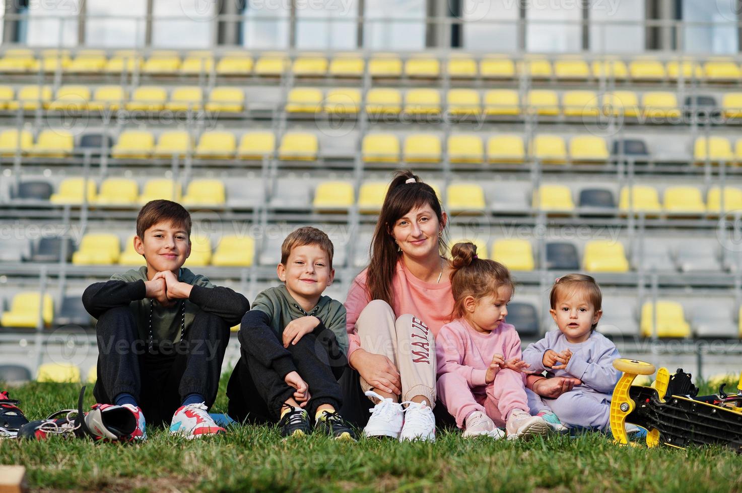 Young stylish mother with four kids sitting on grass against stadium. Sports family spend free time outdoors with scooters and skates. photo