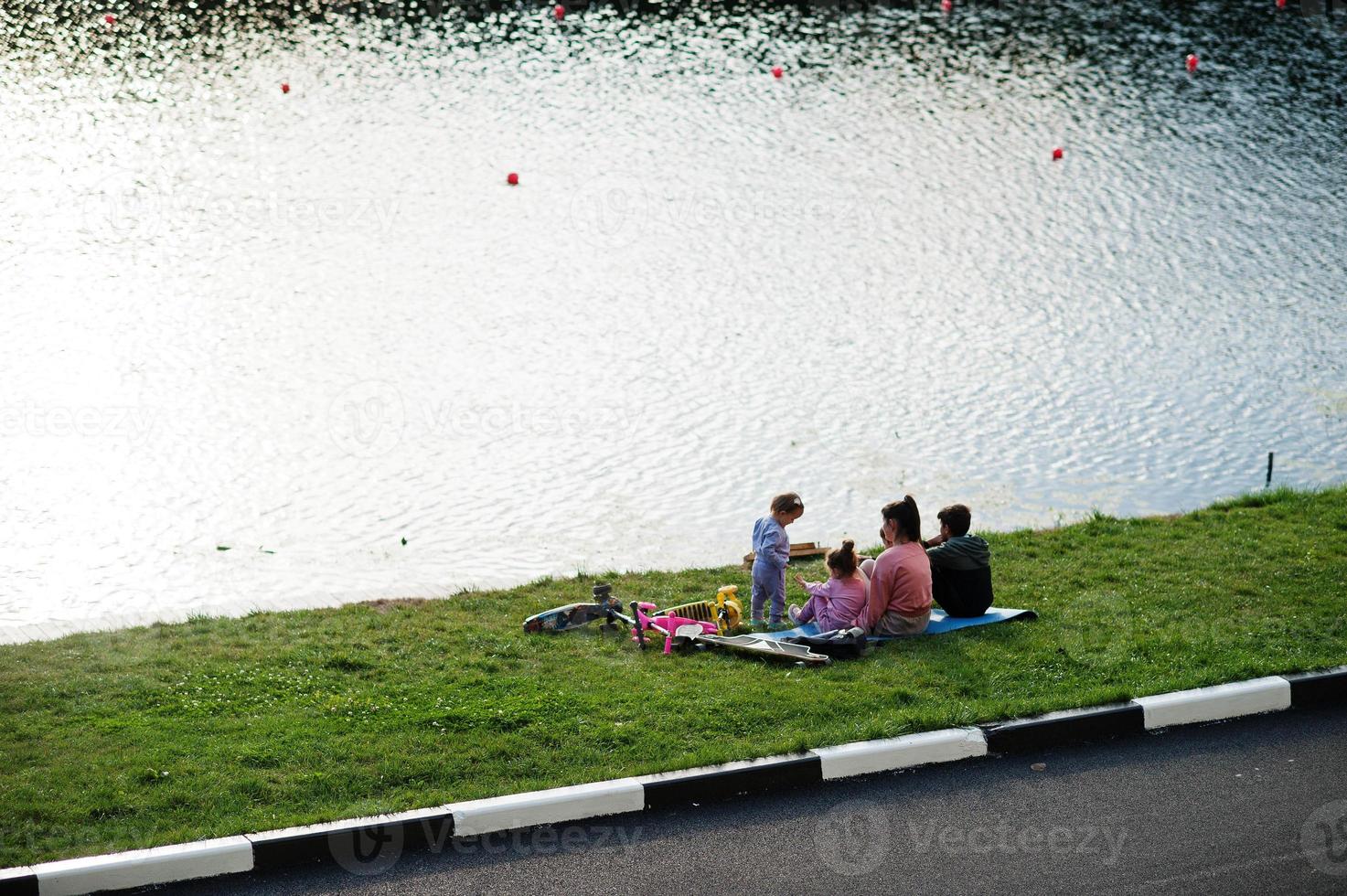 Young stylish mother with four kids sitting on grass against lake. Sports family spend free time outdoors with scooters and skates. photo