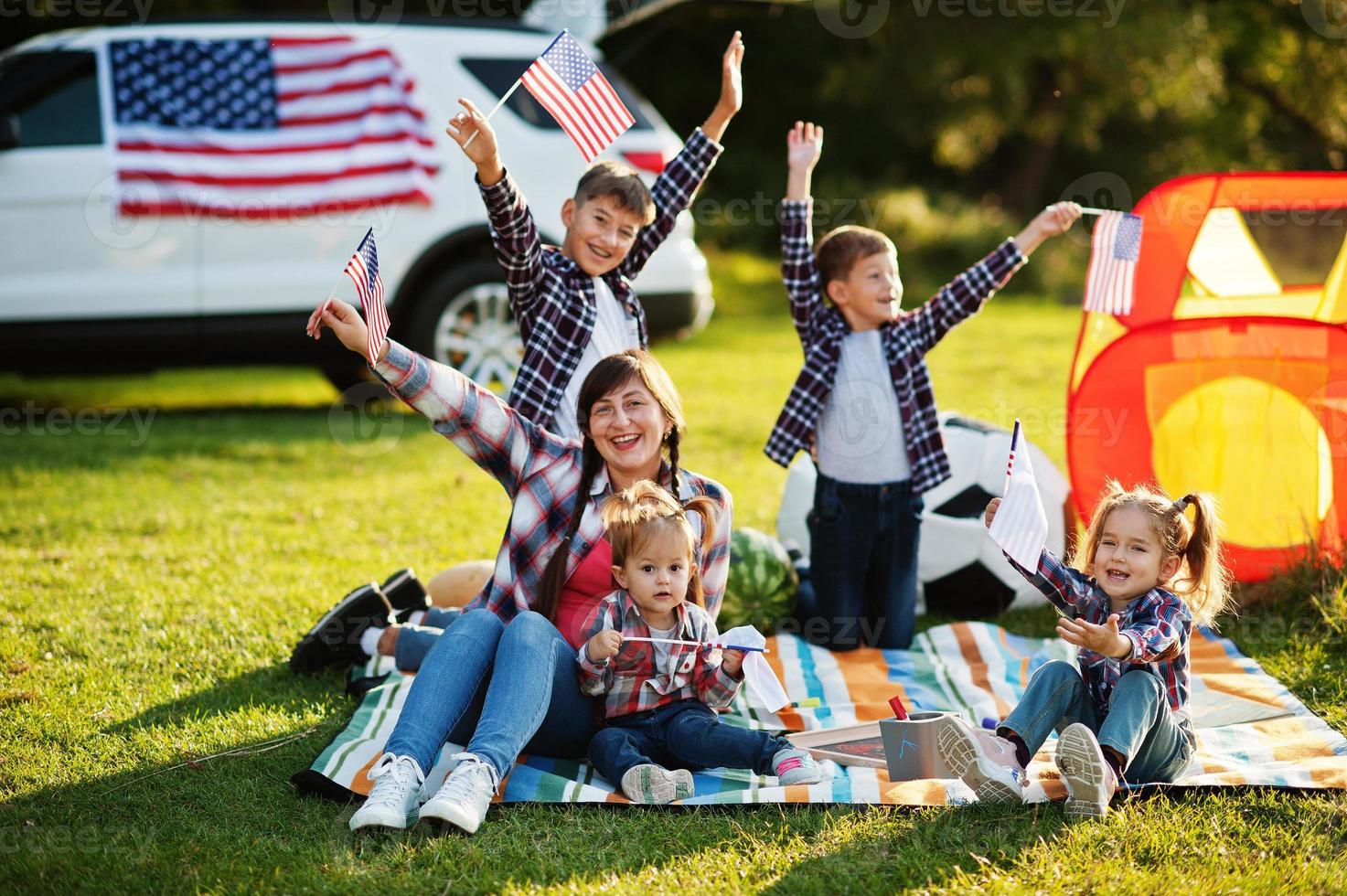 familia americana pasando tiempo juntos. con banderas de estados unidos contra un gran coche suv al aire libre. foto