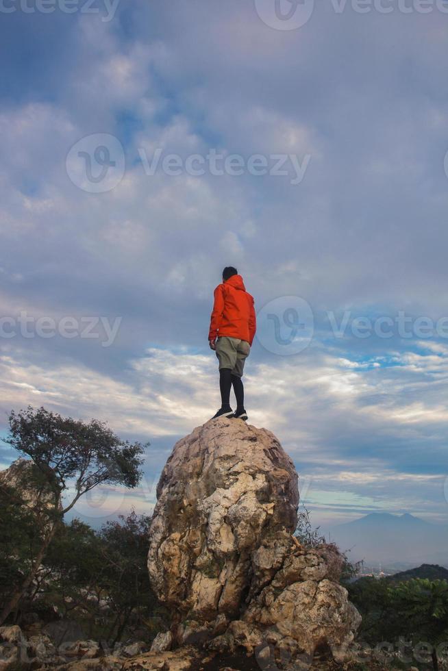 hombre excursionista con chaqueta naranja de pie sobre la piedra y disfrutando de una vista increíble del cielo azul brillante. concepto de viaje y estilo de vida activo. foto