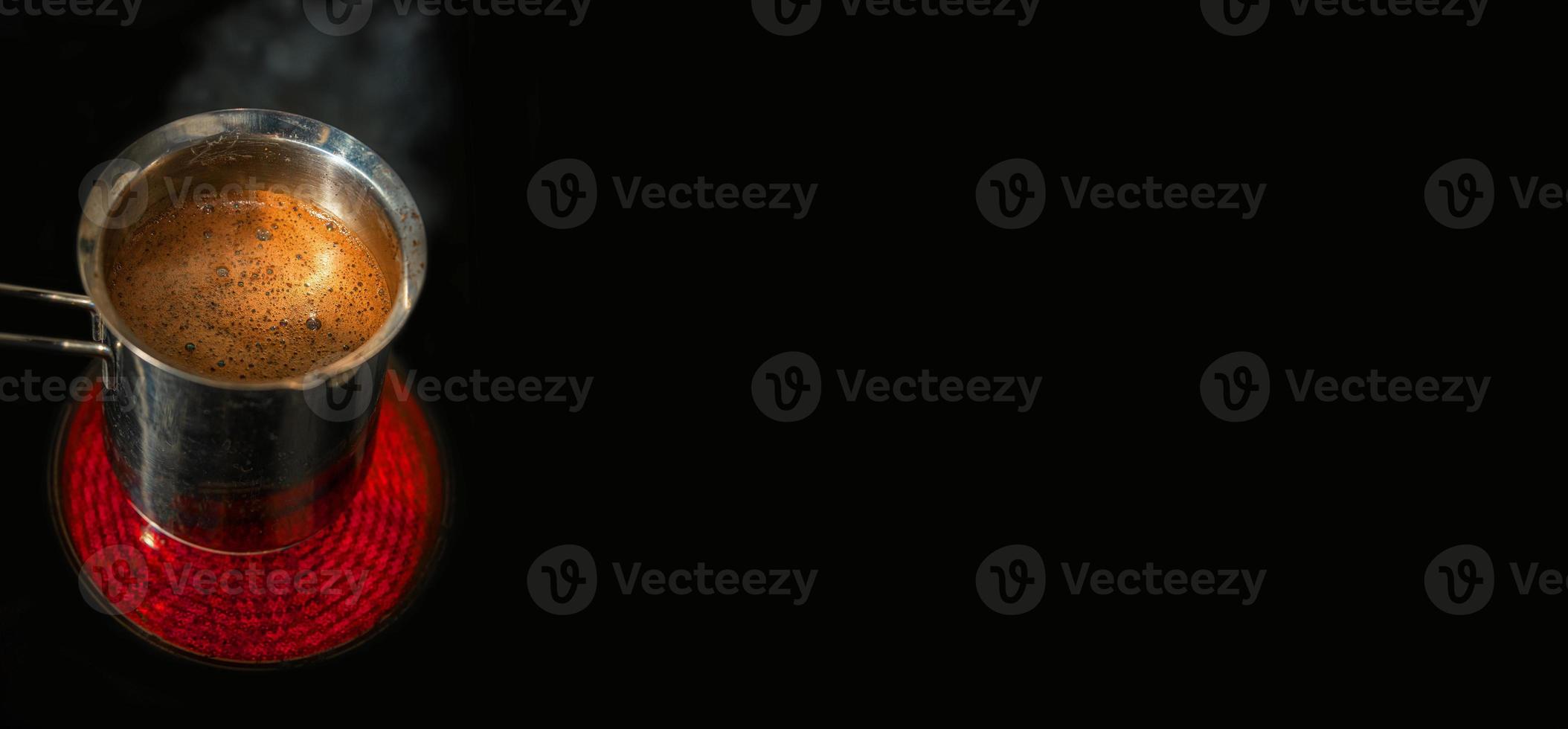 Banner with stainless steel cezve with coffee in process of making standing at hot and red electrical stove in darkness at black solid background with copy space. photo