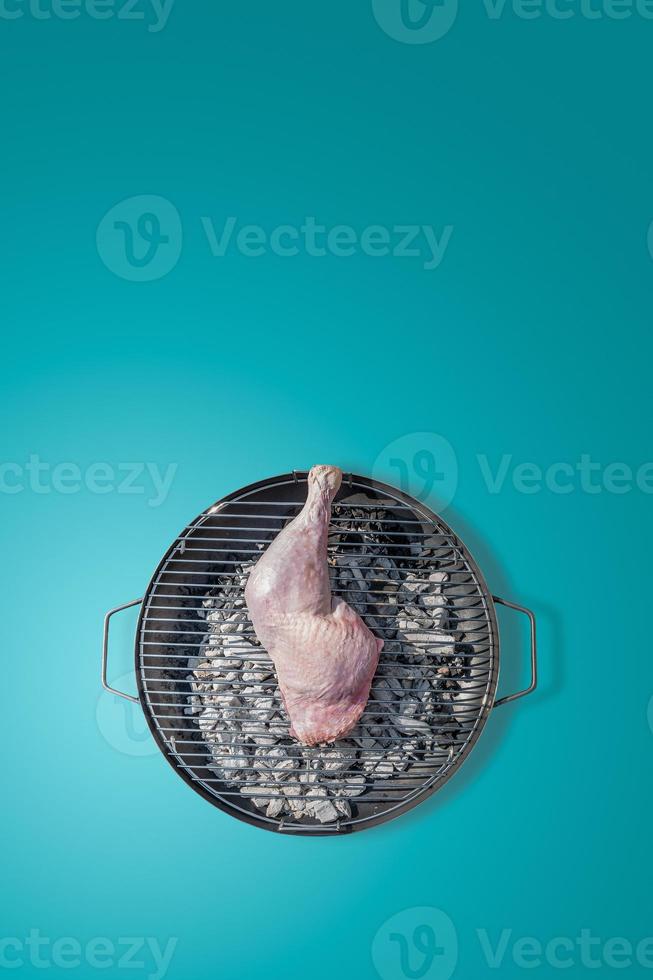 Cover page with big fresh chicken leg prepared for grill at a round barbeque firepan isolated at turquoise gradient background with copy space. Concept of food in outdoor cooking. photo