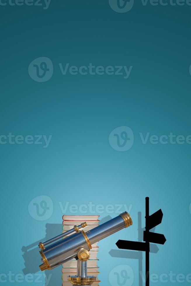 Cover page with old antique metal telescope standing near stack of books with a post showing directions at gradient blue background with copy space and shadow. Concept exploration, education, travel. photo