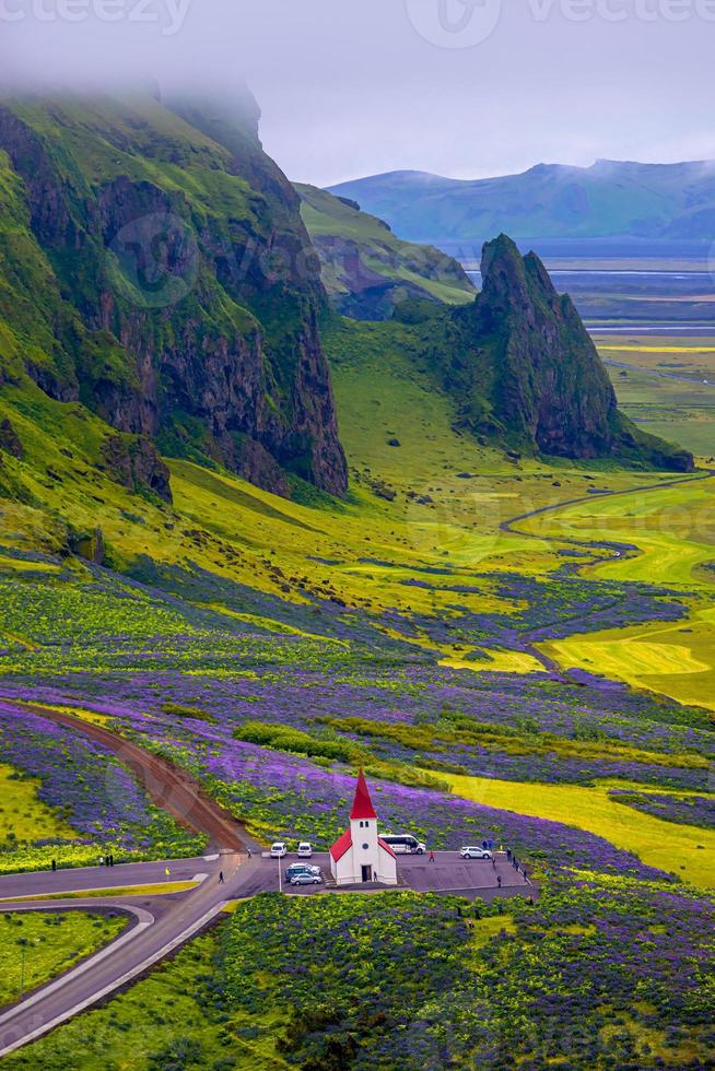 Lonely Lutheran Myrdal church surrounded by violet and pink lupine and yellow meadow flowers at Vik town, South Iceland, at summer sunny day with many visitors and tourists. photo