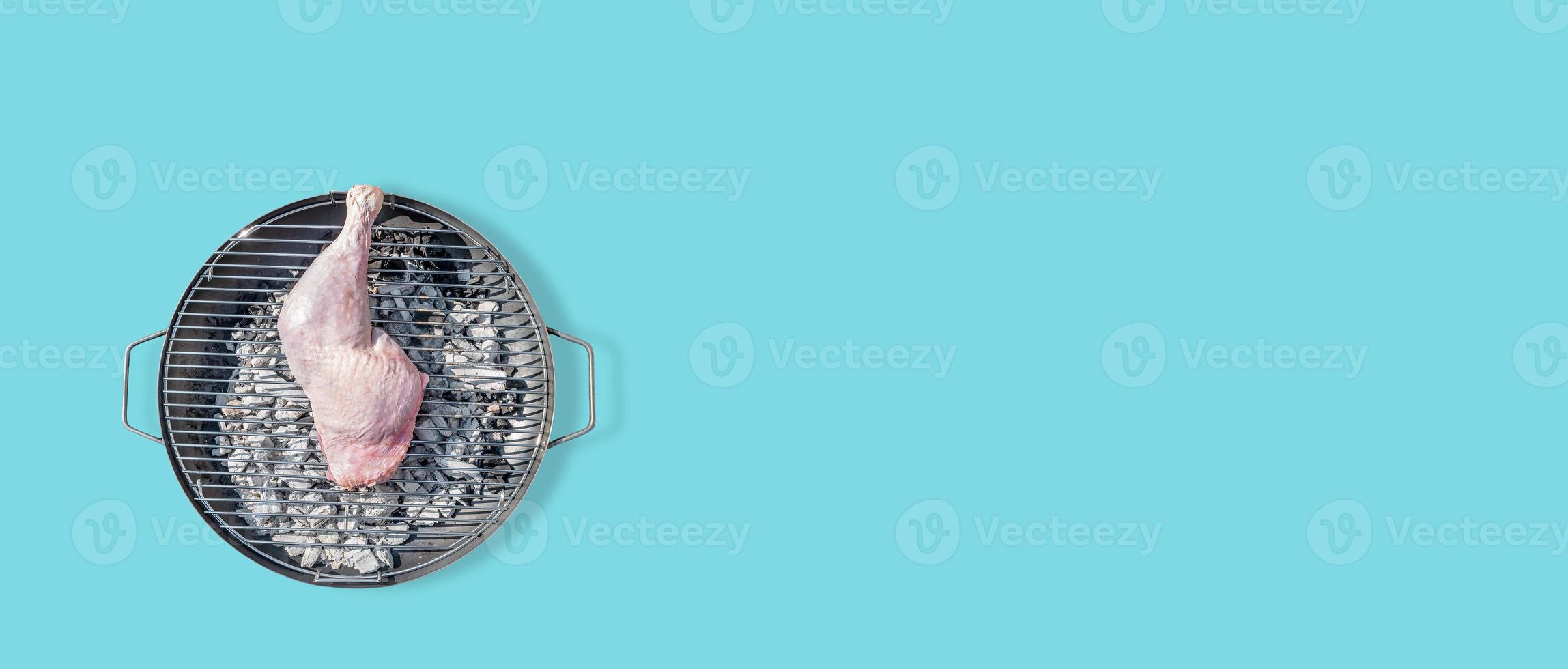Banner with big chicken leg prepared for grill at a round barbeque firepan isolated at turquoise solid background with copy space. Concept of food in outdoor cooking. photo