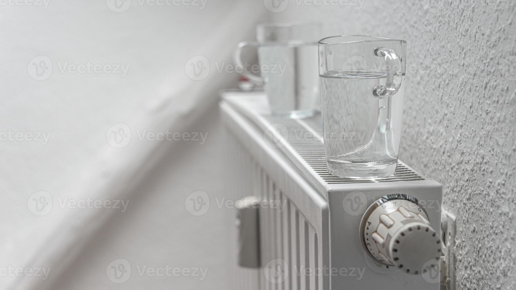 House heating battery with maximum heating power setting and glasses of water standing on it to decrease air dryness at smooth background with copy space. Concept of comfort of home living lifehacks. photo