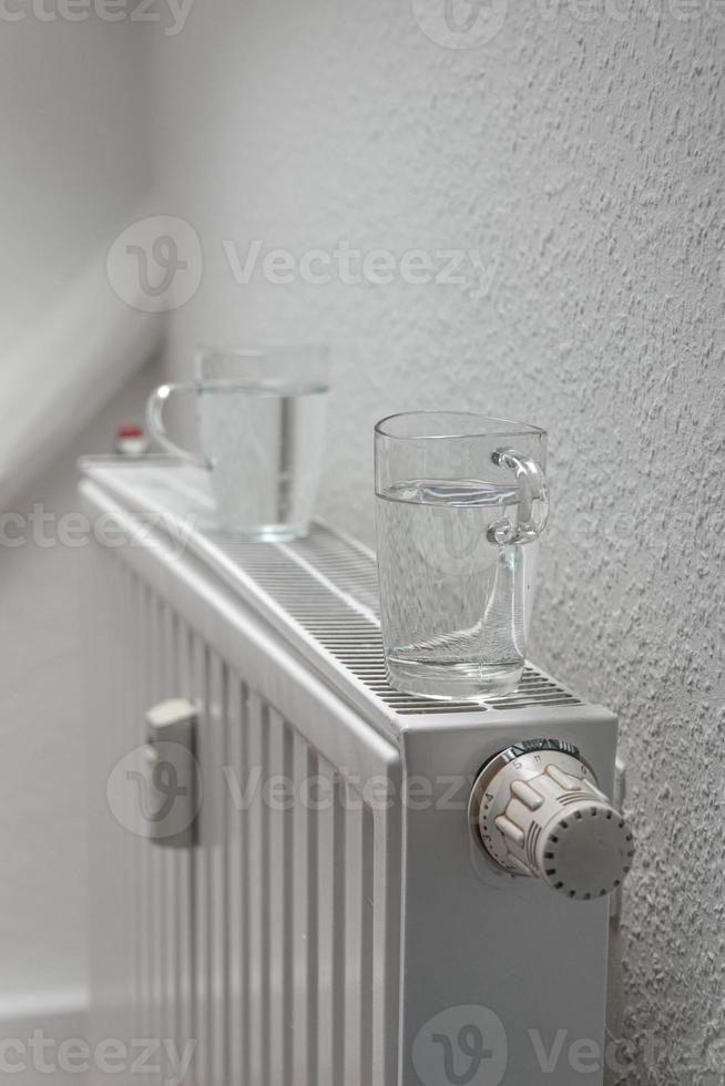 House heating battery with maximum heating power setting and glasses of water standing on it to decrease air dryness at smooth background with copy space. Concept of comfort of home living lifehacks. photo