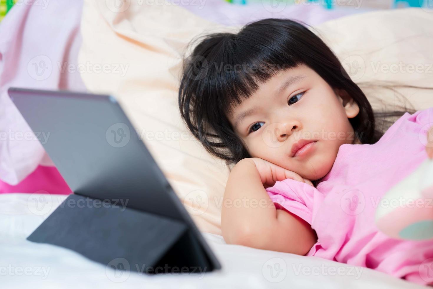 Asian child girl lying down on bed. Kid watching cartoon on touchpad. Children learning online. Upset baby aged 4 years old wearing pink t shirt. photo