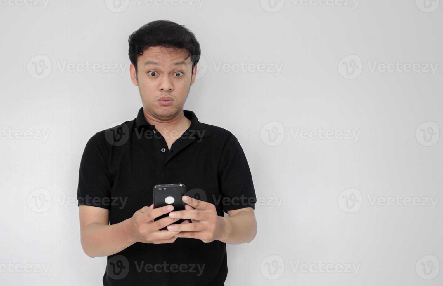 wow, la cara de tu hombre asiático se sorprendió por lo que vio en el teléfono inteligente con un fondo gris aislado. indonesia hombre usar camisa negra aislado fondo gris foto