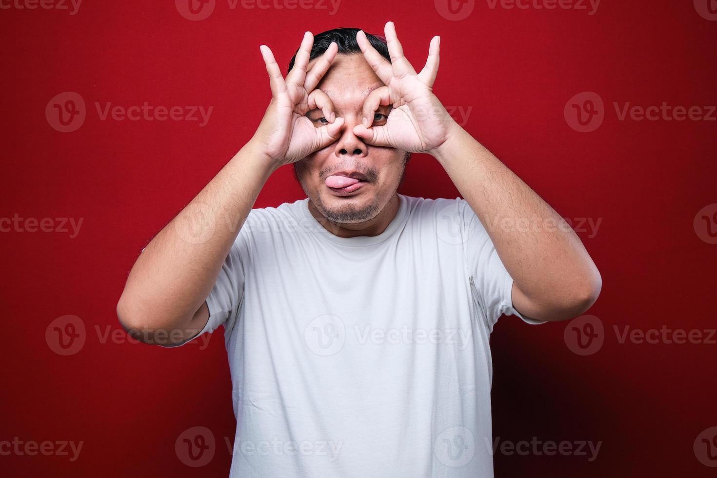Young handsome man wearing white t-shirt doing ok gesture like binoculars sticking tongue out, eyes looking through fingers. crazy expression photo