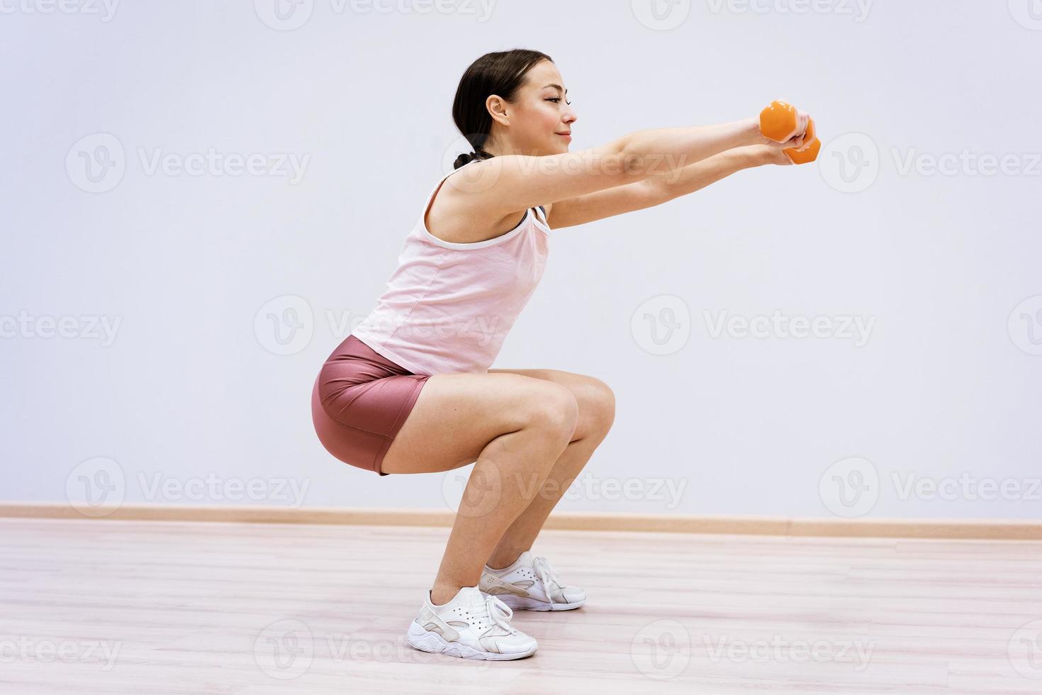 Woman squats with dumbbells in her hand against the background of a light wall photo