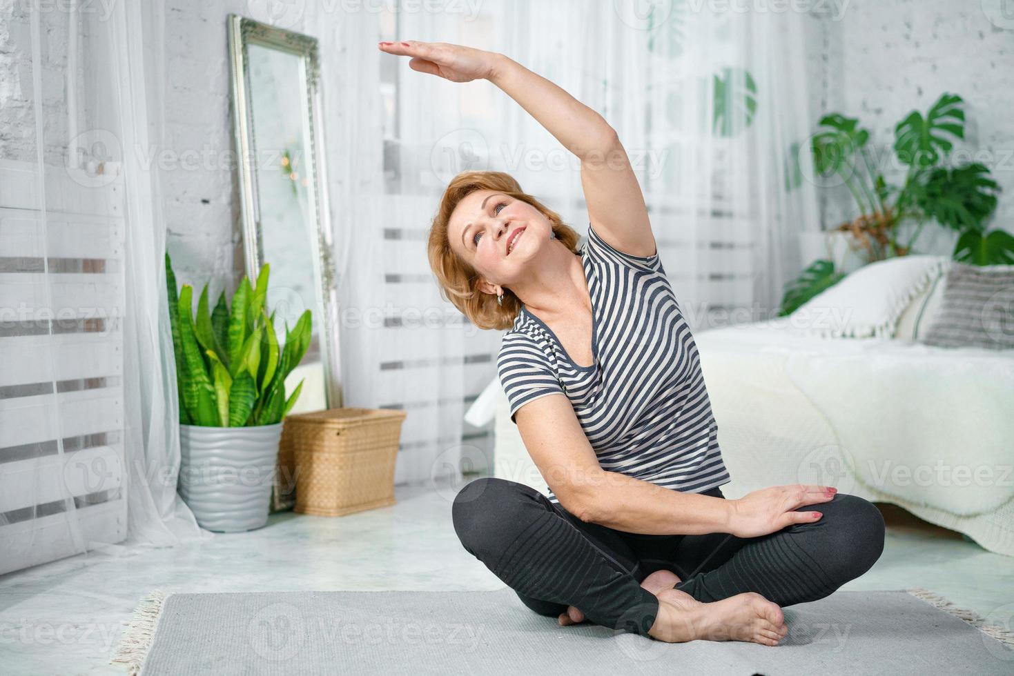 Senior woman exercising while sitting in lotus position. Active mature woman doing stretching exercise in living room at home. photo