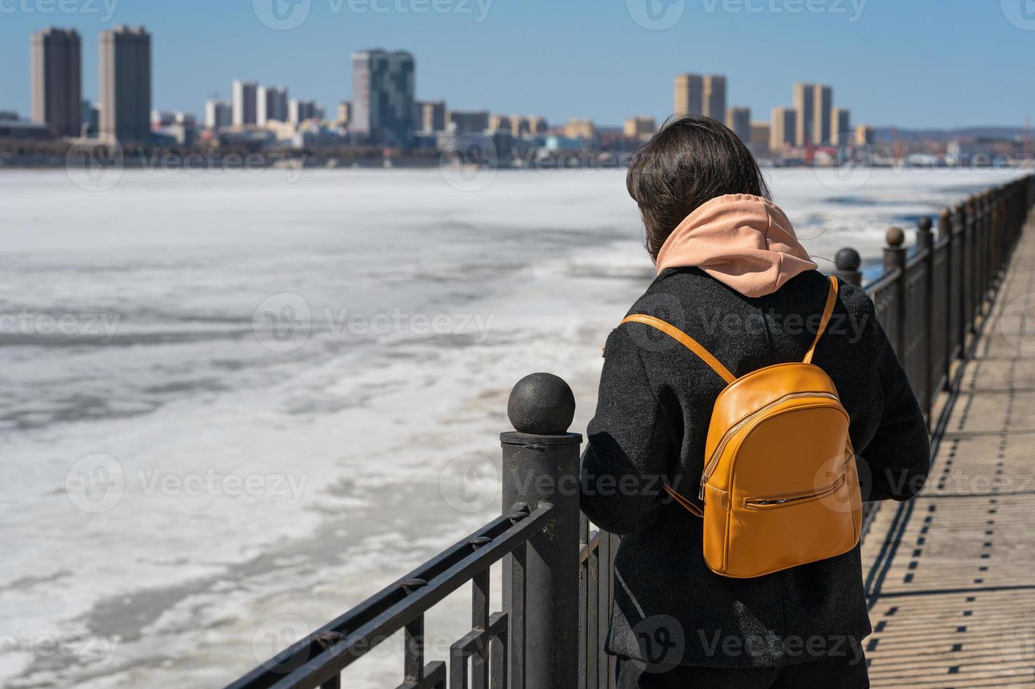 una niña se para en el terraplén y mira la ciudad al otro lado y el hielo en el río foto