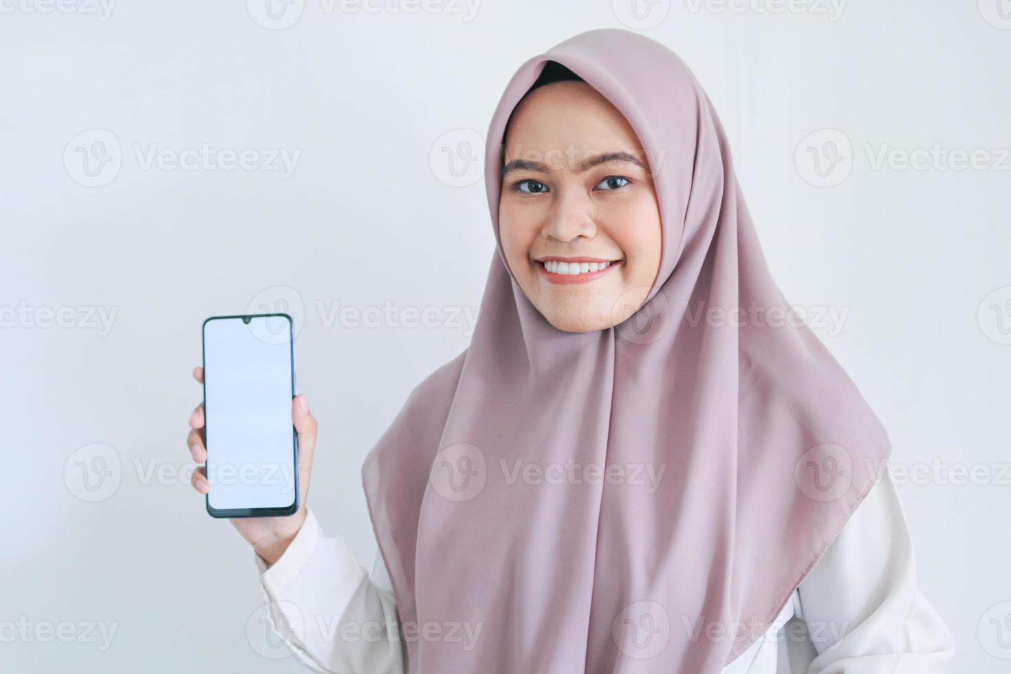 Young Asian Islam woman wearing headscarf is pointing finger on white screen of phone with smile and happy feeling. Indonesian woman on gray background photo