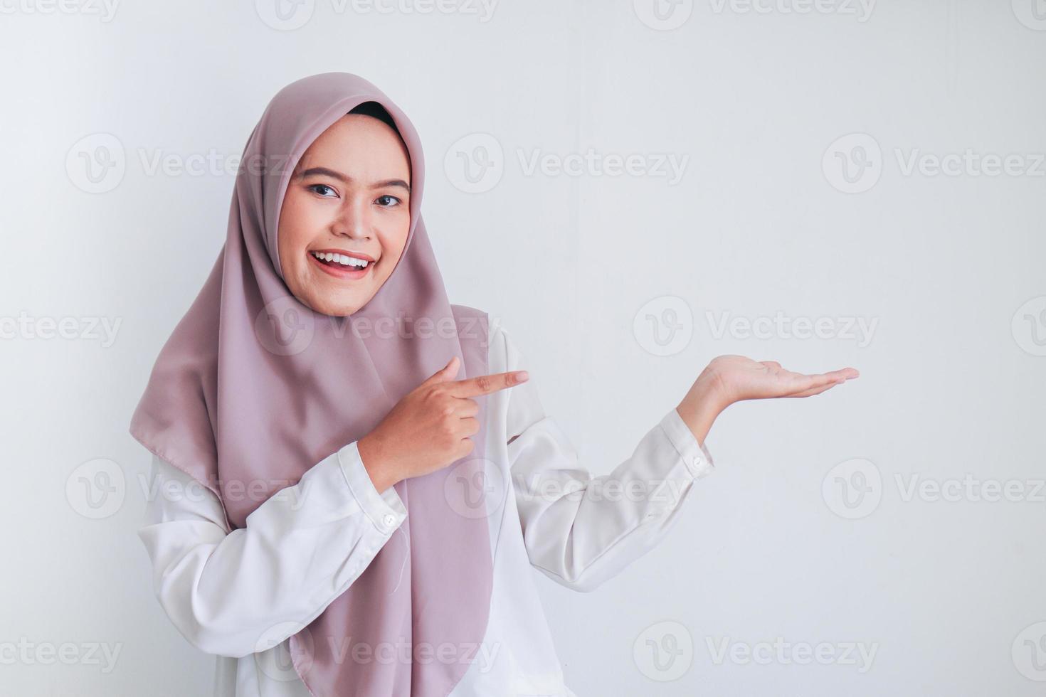 Asian Islam Muslim woman wearing headscarf is pointing finger to blank copy space smile face area. Indonesian woman. Religion concept isolated on gray background photo