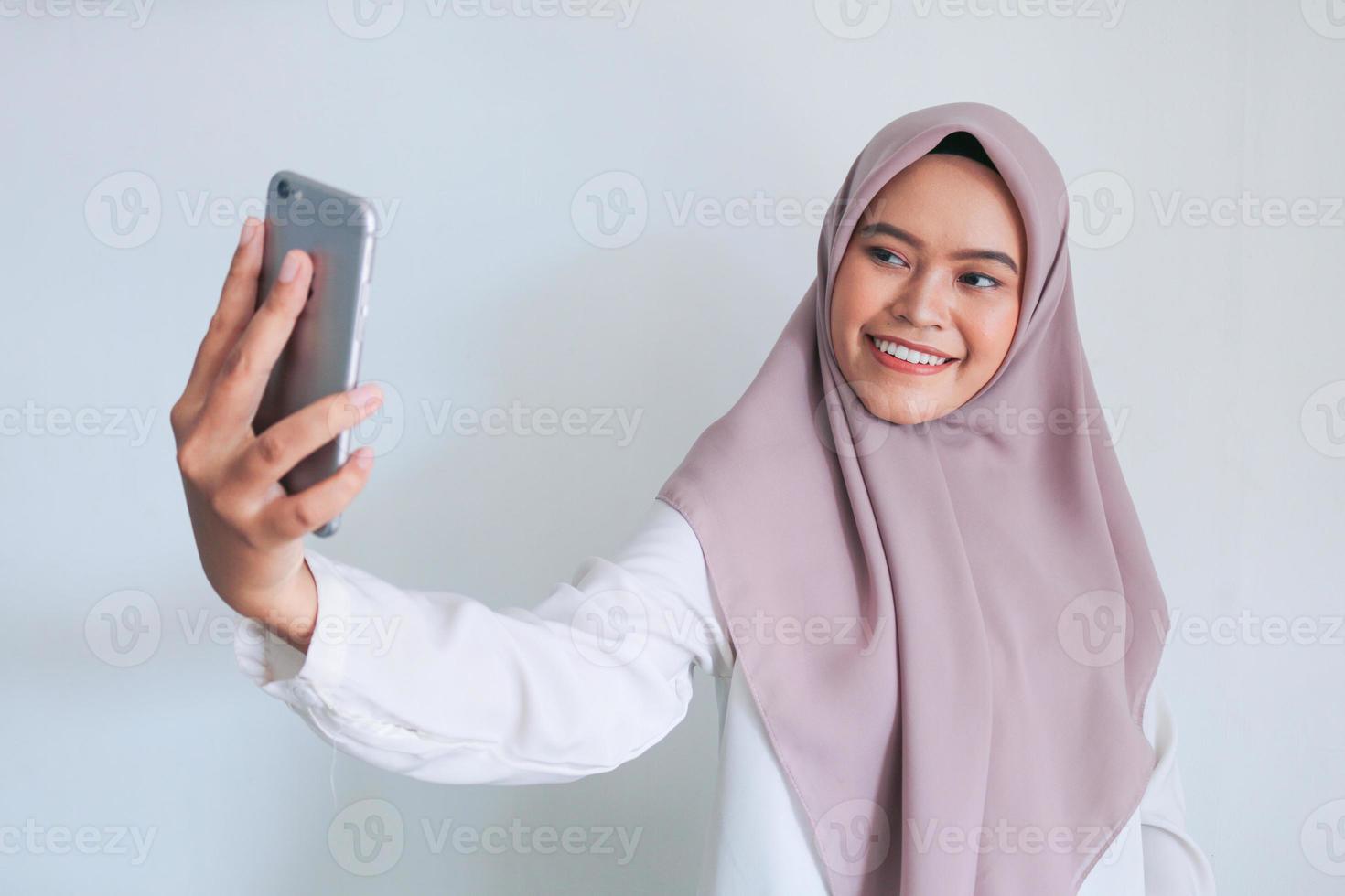 Young Asian Islam woman wearing headscarf is smiling and selfie taking pictures with her mobile phone. Indonesian woman on gray background photo