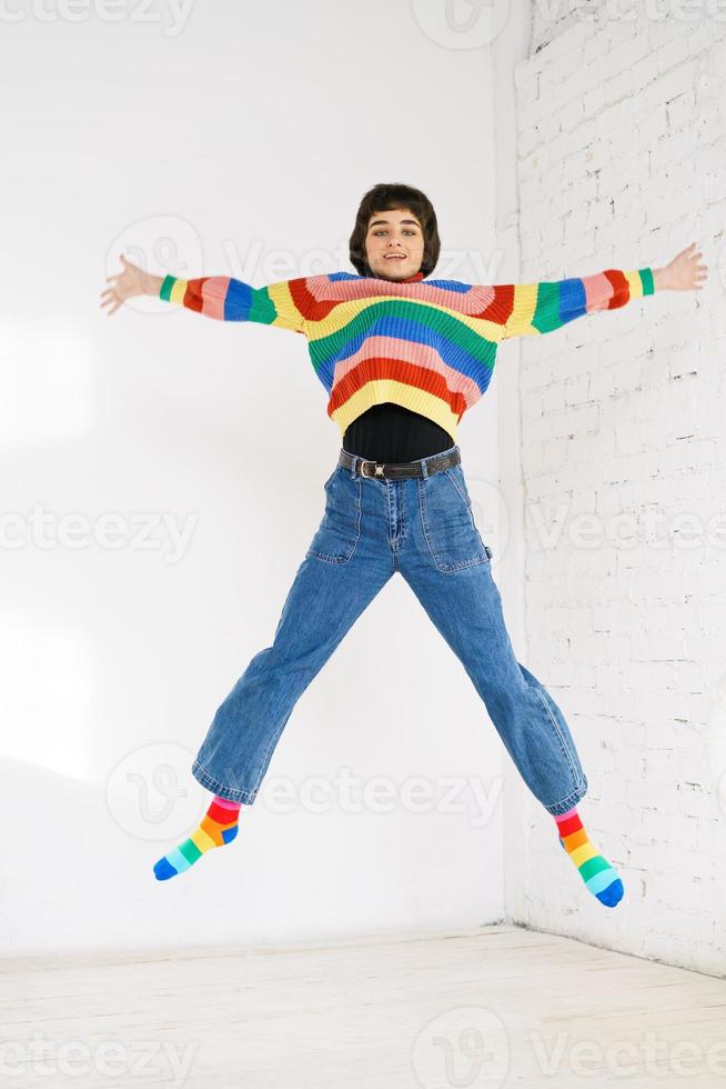 Happy girl in rainbow sweater jumps up on light background. photo