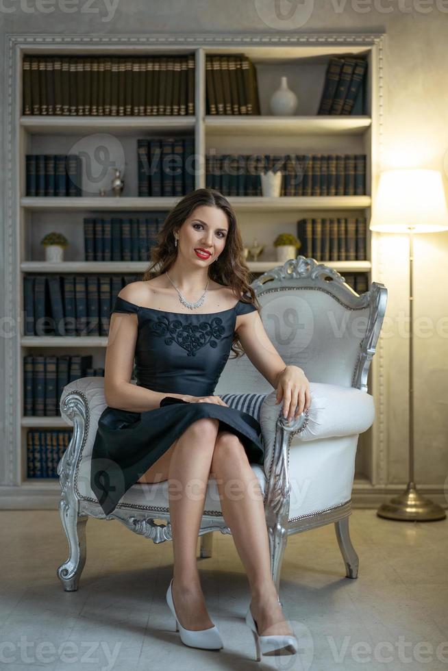 A beautiful woman with makeup and red lipstick is sitting in a black dress on a white chair against the background of shelves with books photo