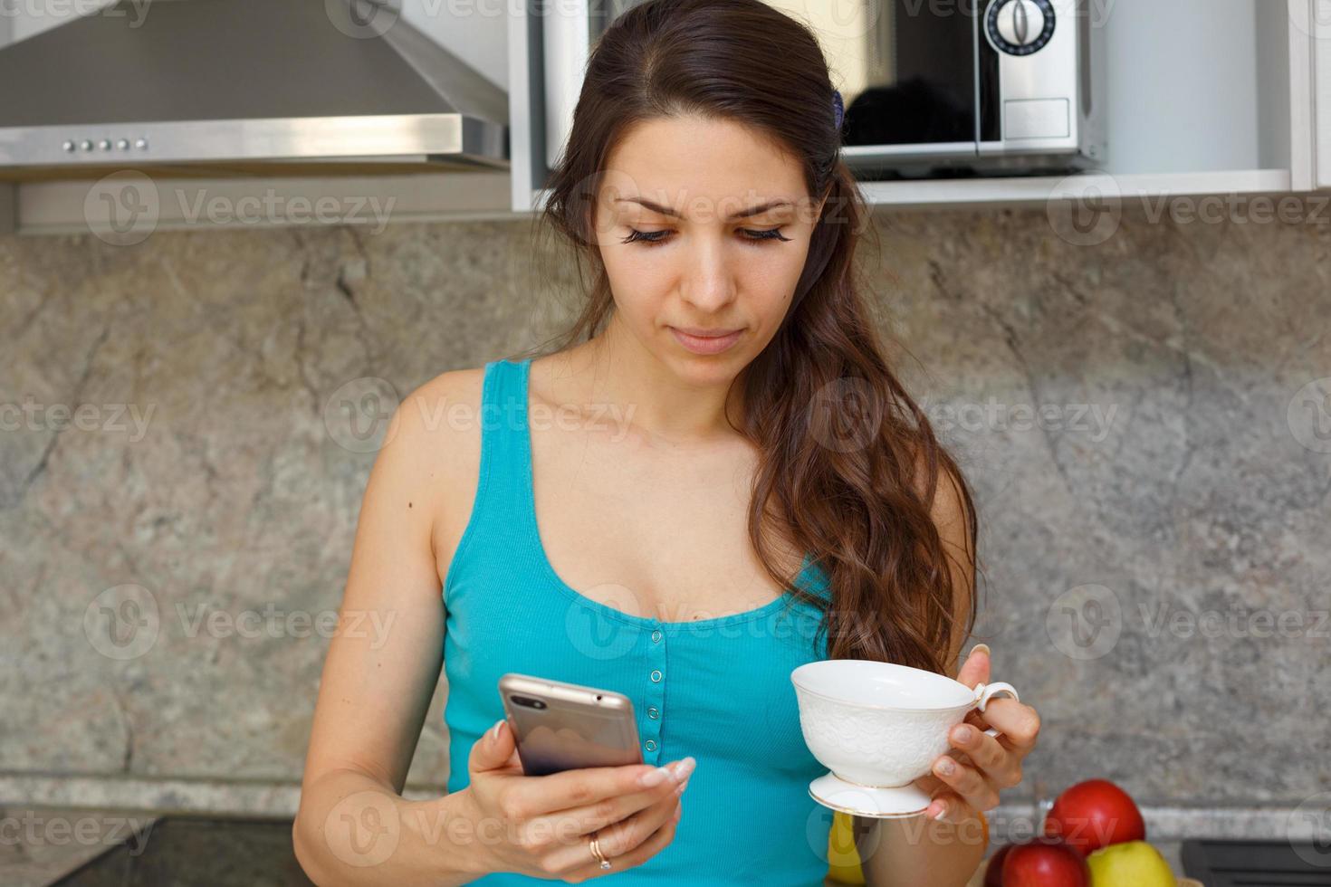a woman in the kitchen stands with a mug and a phone in her hand. photo