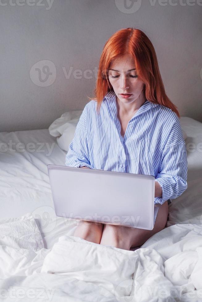 young woman sitting in bed using laptop on her knees doing remote work from home photo