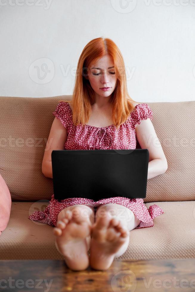 young woman using laptop working from home sitting on couch with feet on table photo