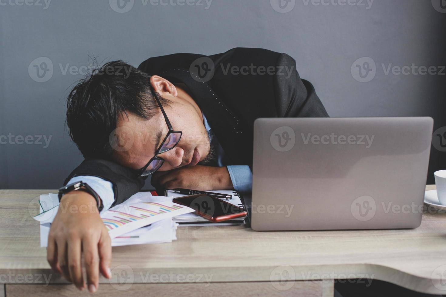 Asian businessman falling asleep at office desk with closed eyes on grey background, overworked young man, unmotivated worker sleeping at workplace, boring of routine work photo