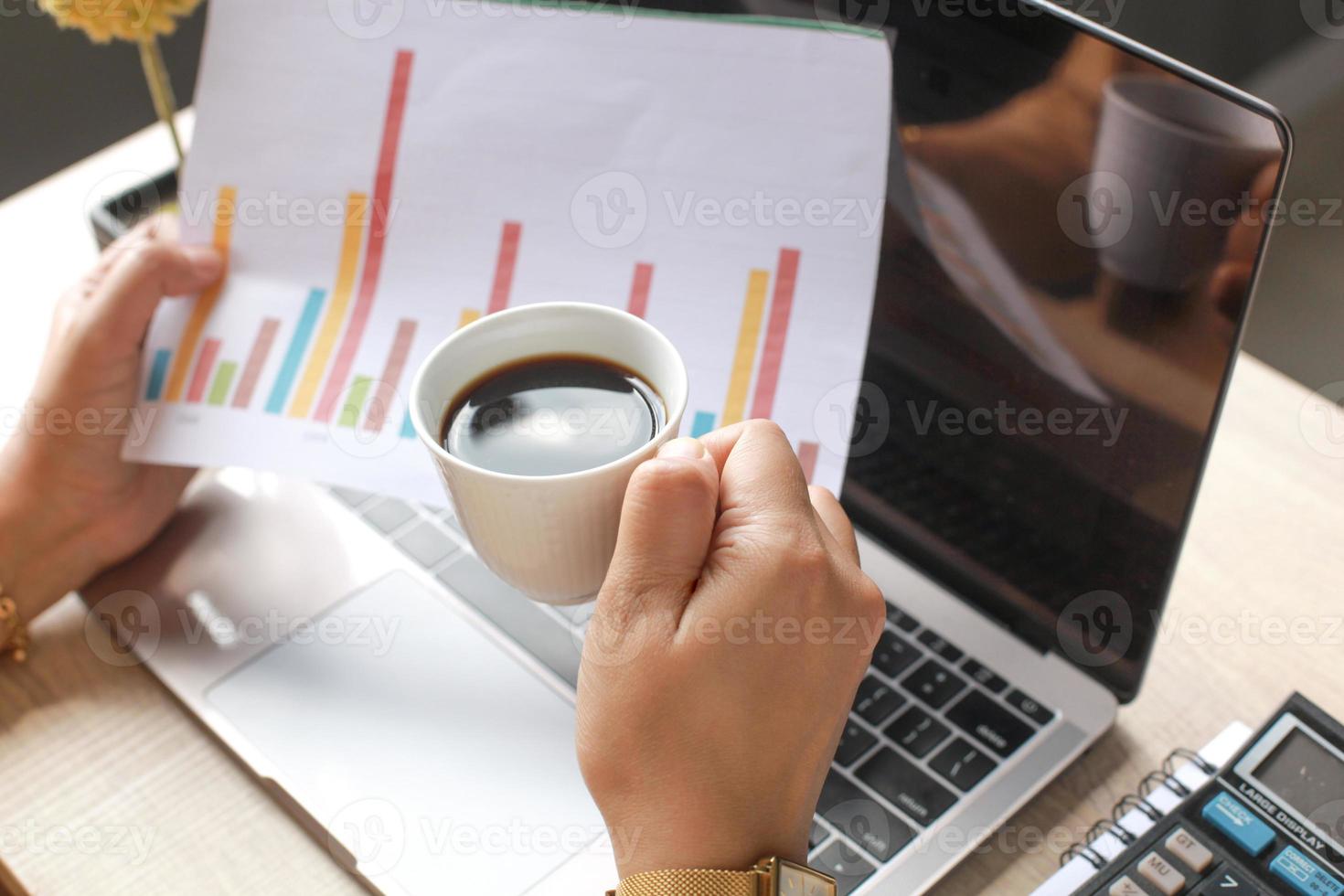 A businessman hand holds a cup of coffee and business reports with laptop, calculator and pen on the book, classic alarm clock, book on wooden office desk photo