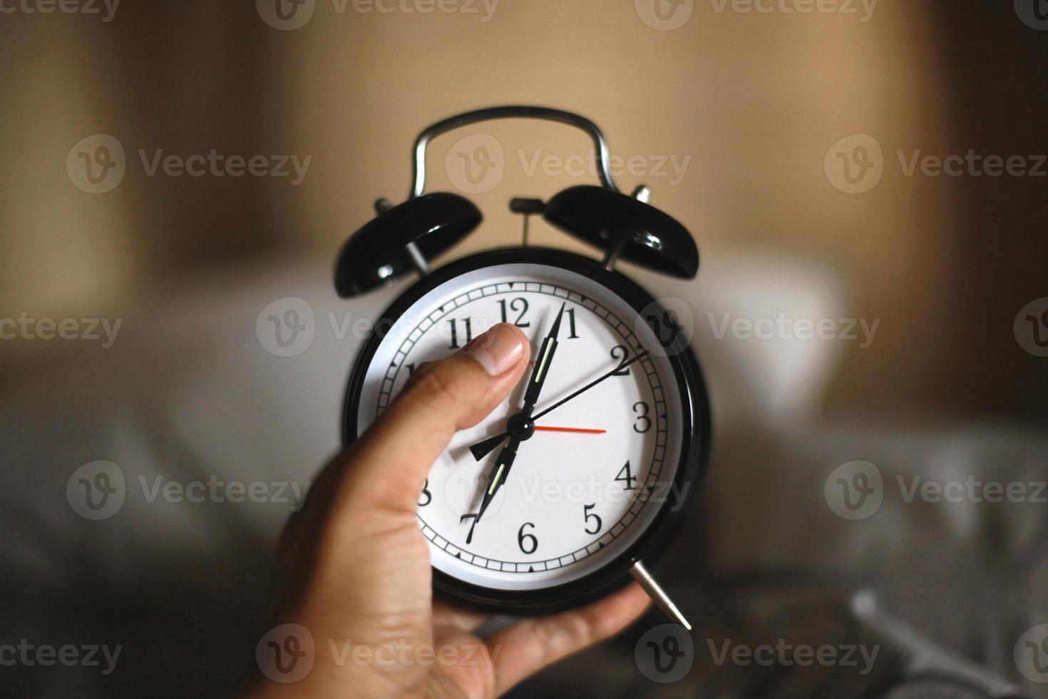 Man hand holding classic black alarm clock showing seven o'clock with blurry background, waking up in the morning concept photo