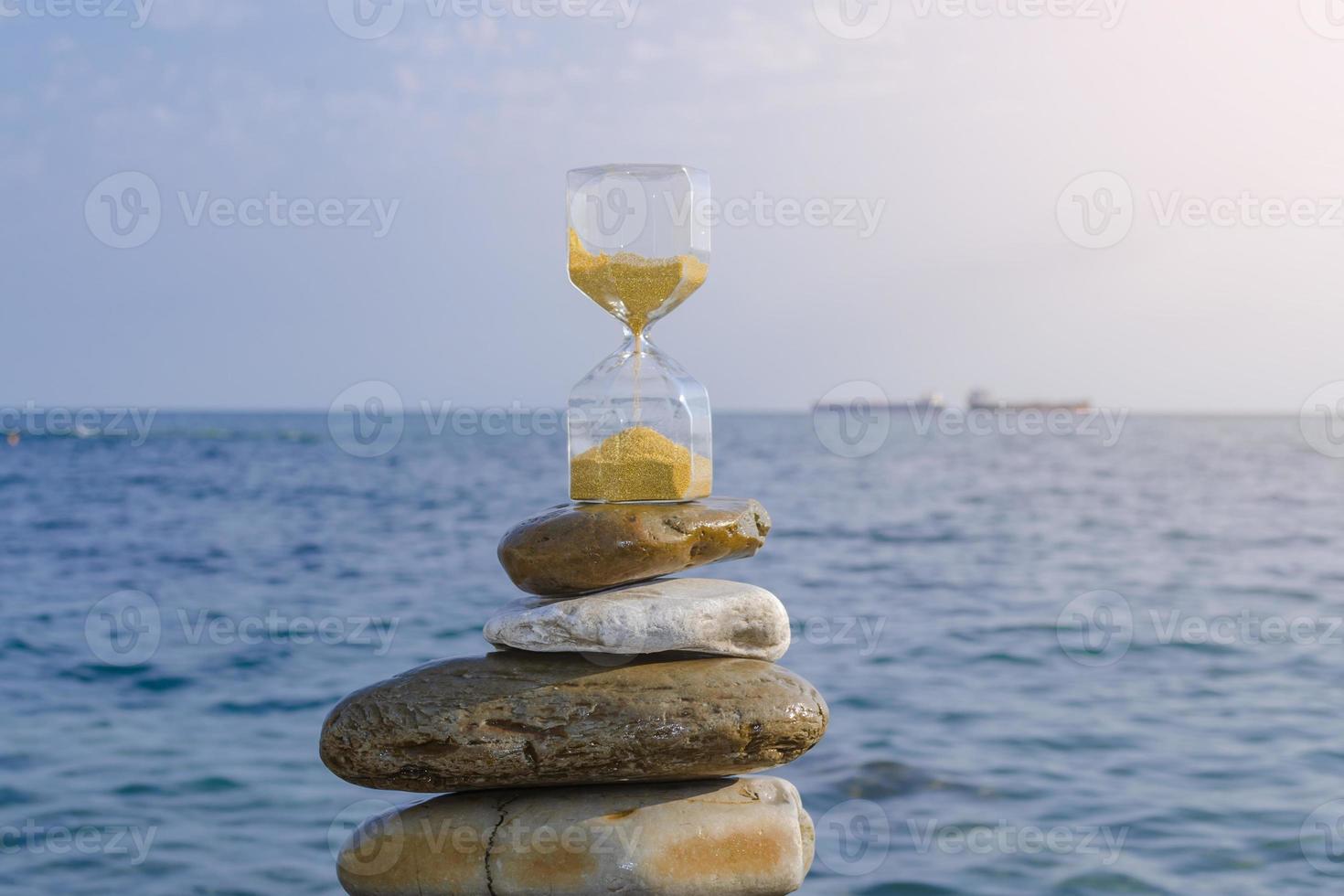 An hourglass with golden sand on sea rocks against a blue sea background. photo