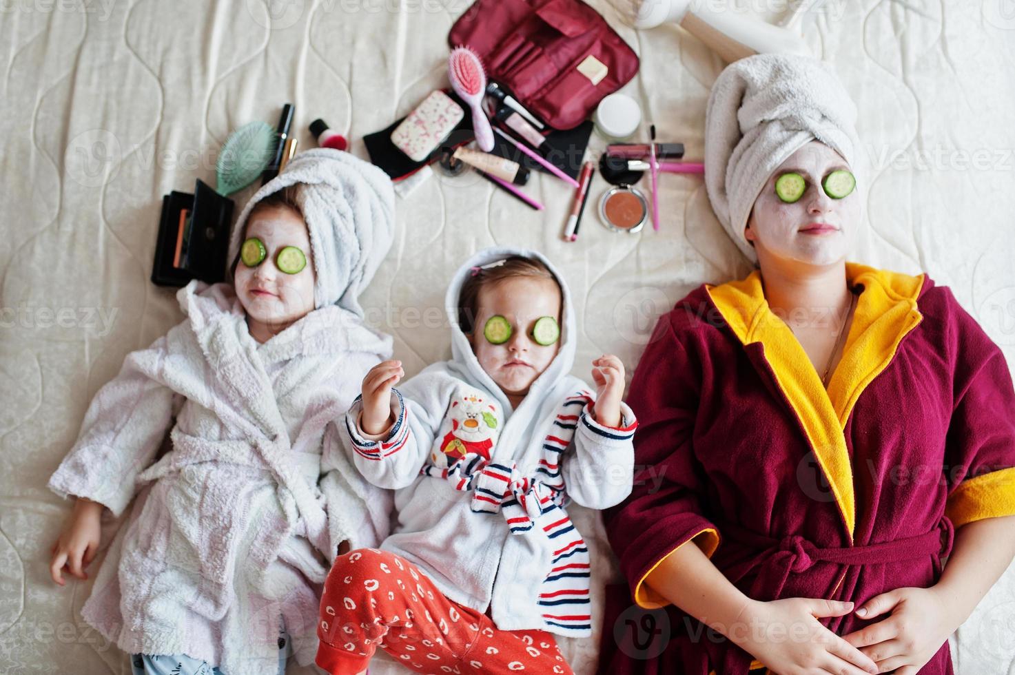 Mother with daughters in a bathrobe with a towel on her head take care of the skin at home, cucumbers on eyes. photo