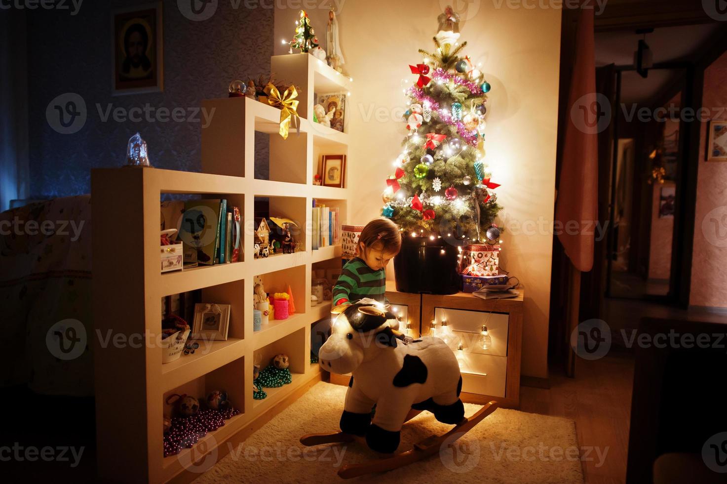 Baby girl on rocking cow toy against Christmas tree with shining garlands on evening home. photo