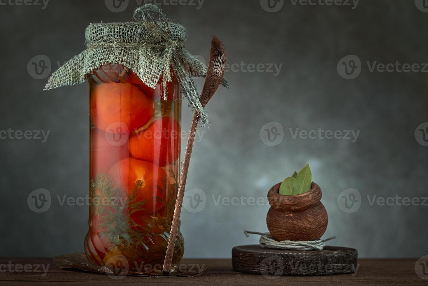 Preserves. Pickled tomato in glass on grey dark background. Next to it is wooden jar with Bay leaf. photo