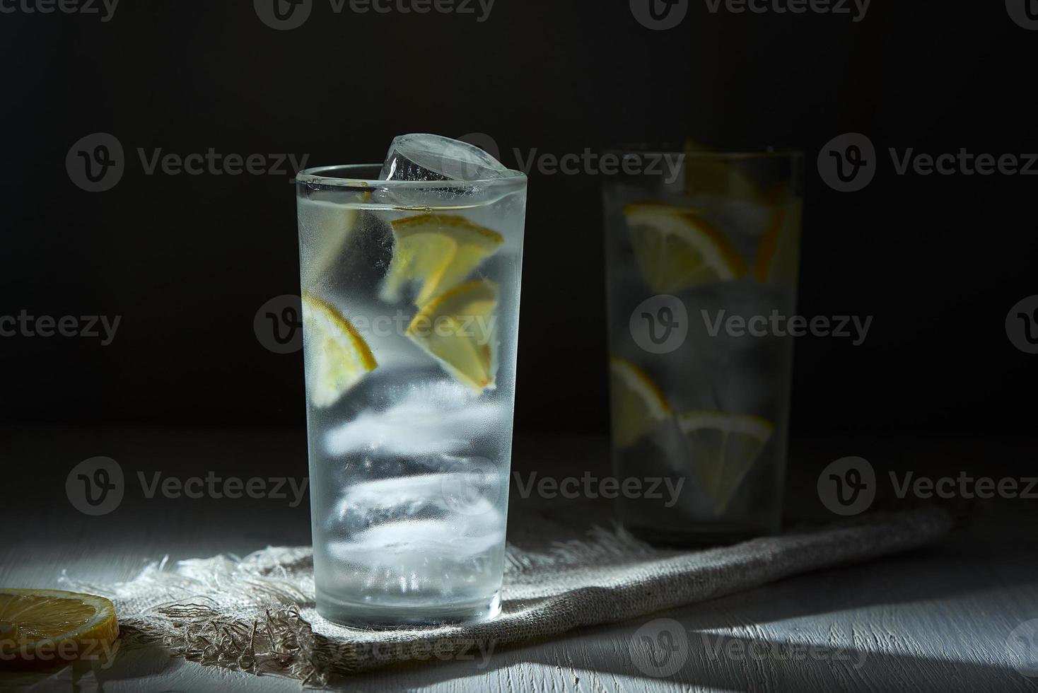 Two glasses of ice cold lemonade with lemon slices and ice on linen napkin on black background in blur. photo