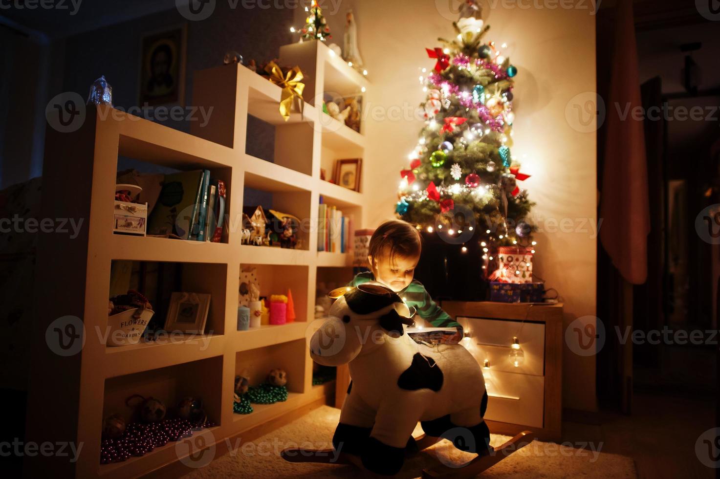 Baby girl on rocking cow toy against Christmas tree with shining garlands on evening home. photo