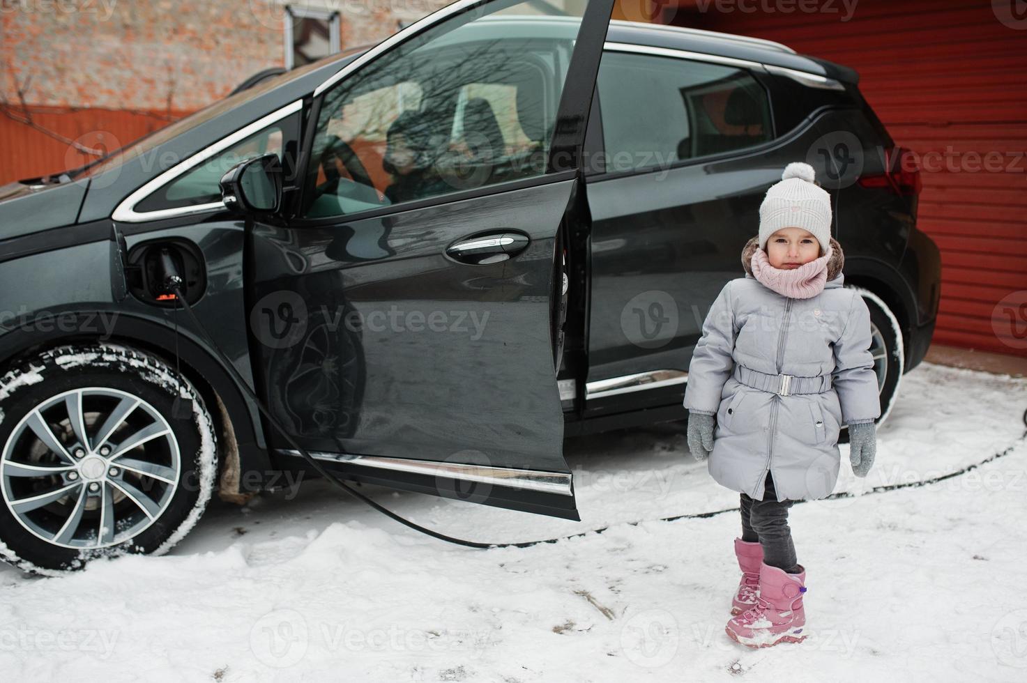 Baby girl against charging electric car in the yard of house at winter. photo