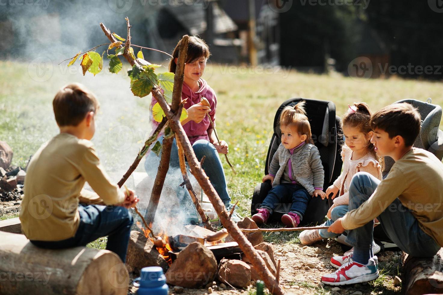 Family bonfire in mountain. Frying sausages. Mother with four kids camping. Autumn hike and camp weather. Warming and cooking near flame together. photo