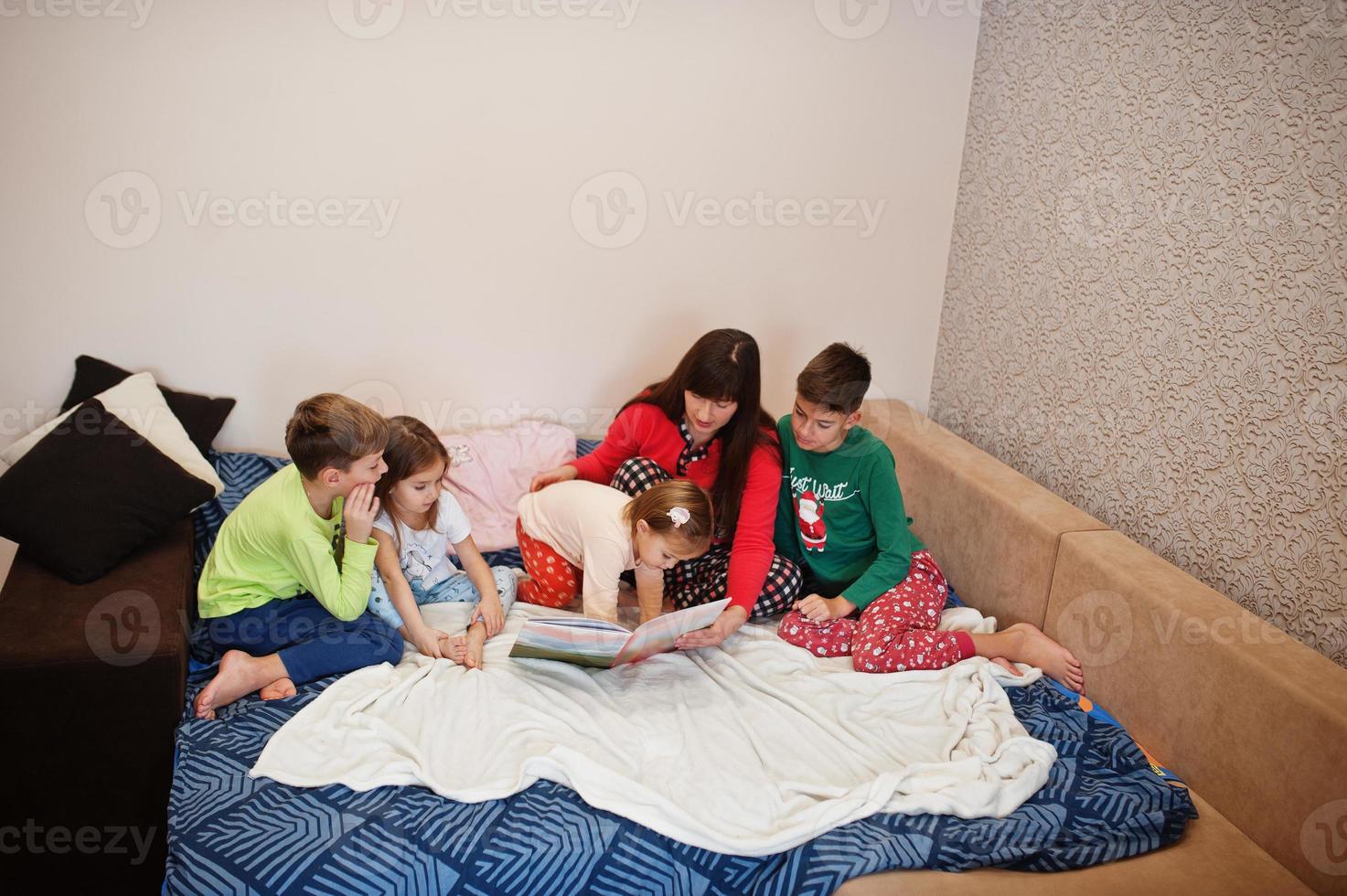 Happy big family is having fun together in bedroom. Large family morning concept. Mother with four kids wear pajamas read book in bed at home. photo