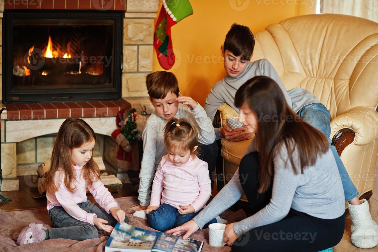 Happy young large family by a fireplace in warm living room on winter day. Mother with four kids at home read book. photo