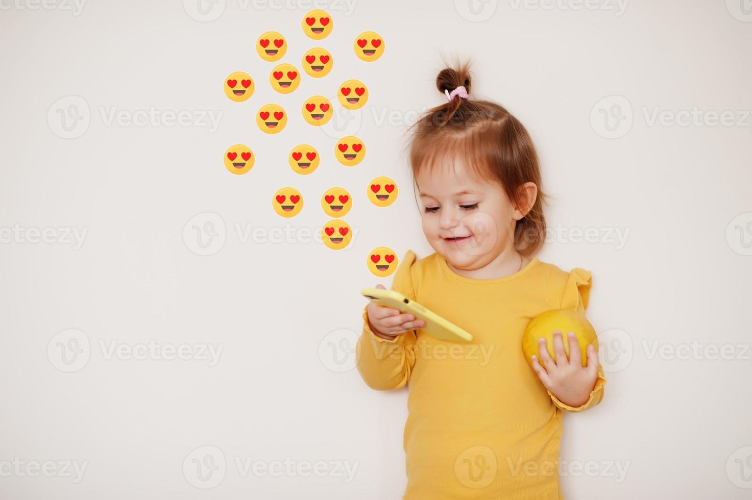 Baby girl in yellow with lemon and mobile phone with heart eyes emoji, isolated background. photo