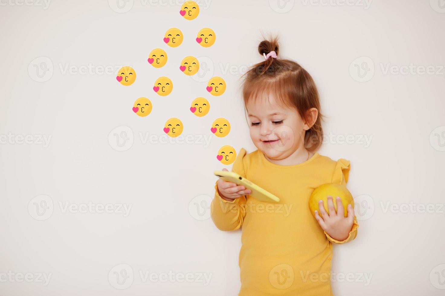 Baby girl in yellow with lemon and mobile phone with kiss emoji, isolated background. photo